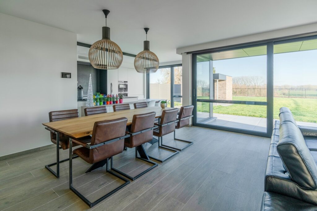 Open leefruimte met eetkamer en keuken, afgewerkt met keramische tegels in houtlook die doorlopen tot aan de grote schuiframen.